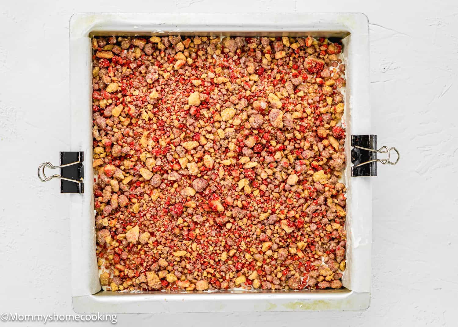A square baking pan filled with an unbaked, crumbly Eggless Strawberry Crunch Cake mixture, lined with parchment paper secured by binder clips.