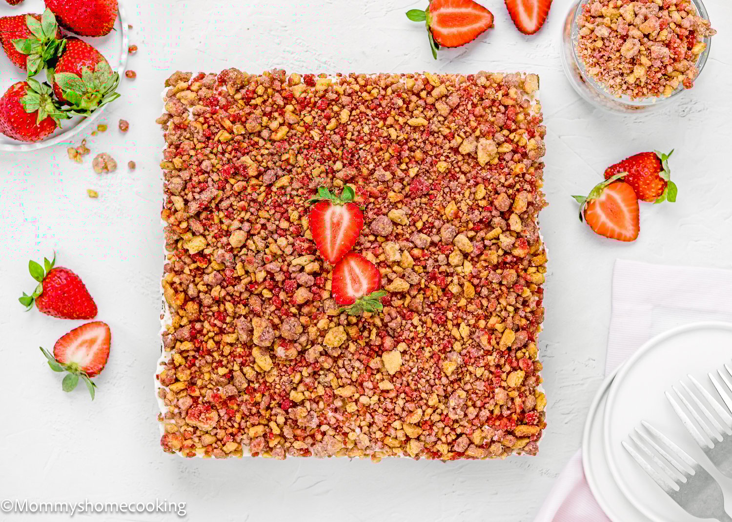 An Eggless Strawberry Crunch Cake, square-shaped and topped with crushed crumbs and halved strawberries, is surrounded by fresh strawberries and utensils on a white surface.