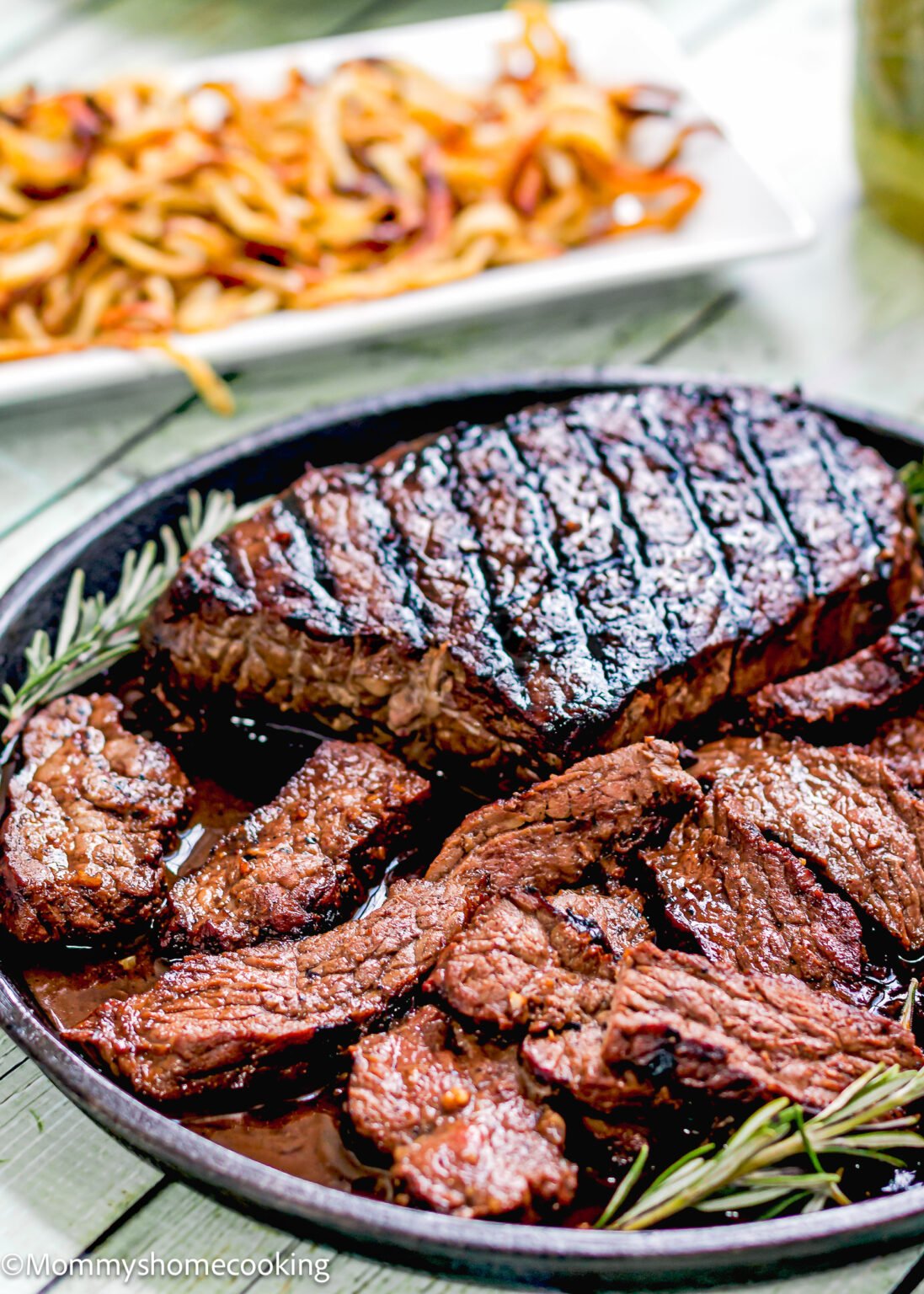 Grilled Balsamic Garlic Steak is sliced and served in a round dish with a rosemary garnish, with a plate of curly fries in the background.