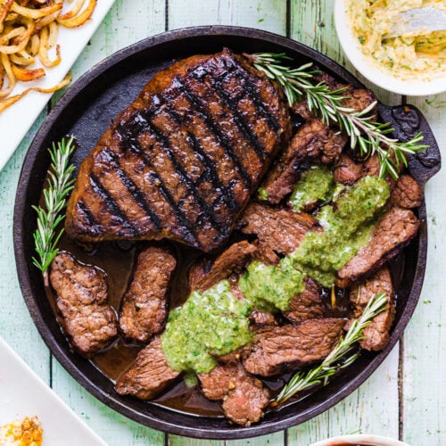 Grilled Balsamic Garlic Steak and sliced beef topped with green sauce, garnished with rosemary on a round plate, served with curly fries, butter, and dipping sauces.
