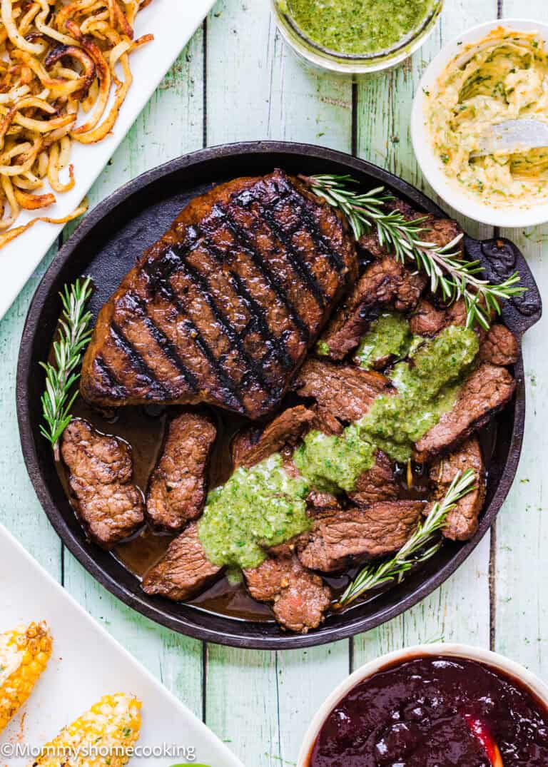 Grilled Balsamic Garlic Steak and sliced beef topped with green sauce, garnished with rosemary on a round plate, served with curly fries, butter, and dipping sauces.