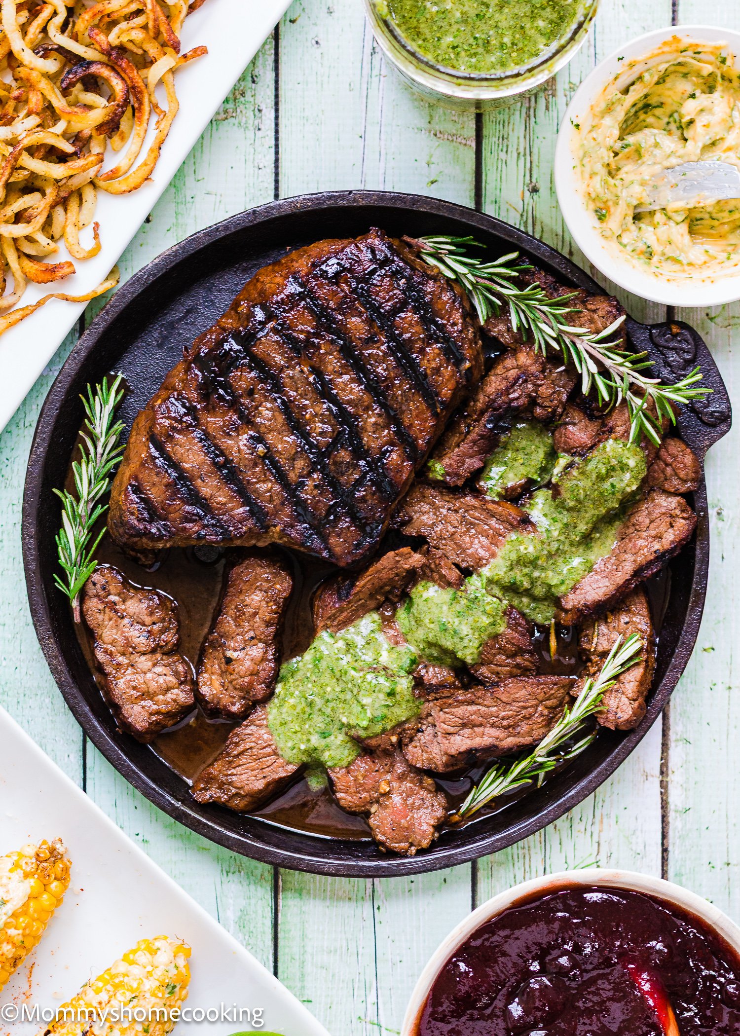 Grilled Balsamic Garlic Steak and sliced beef topped with green sauce, garnished with rosemary on a round plate, served with curly fries, butter, and dipping sauces.