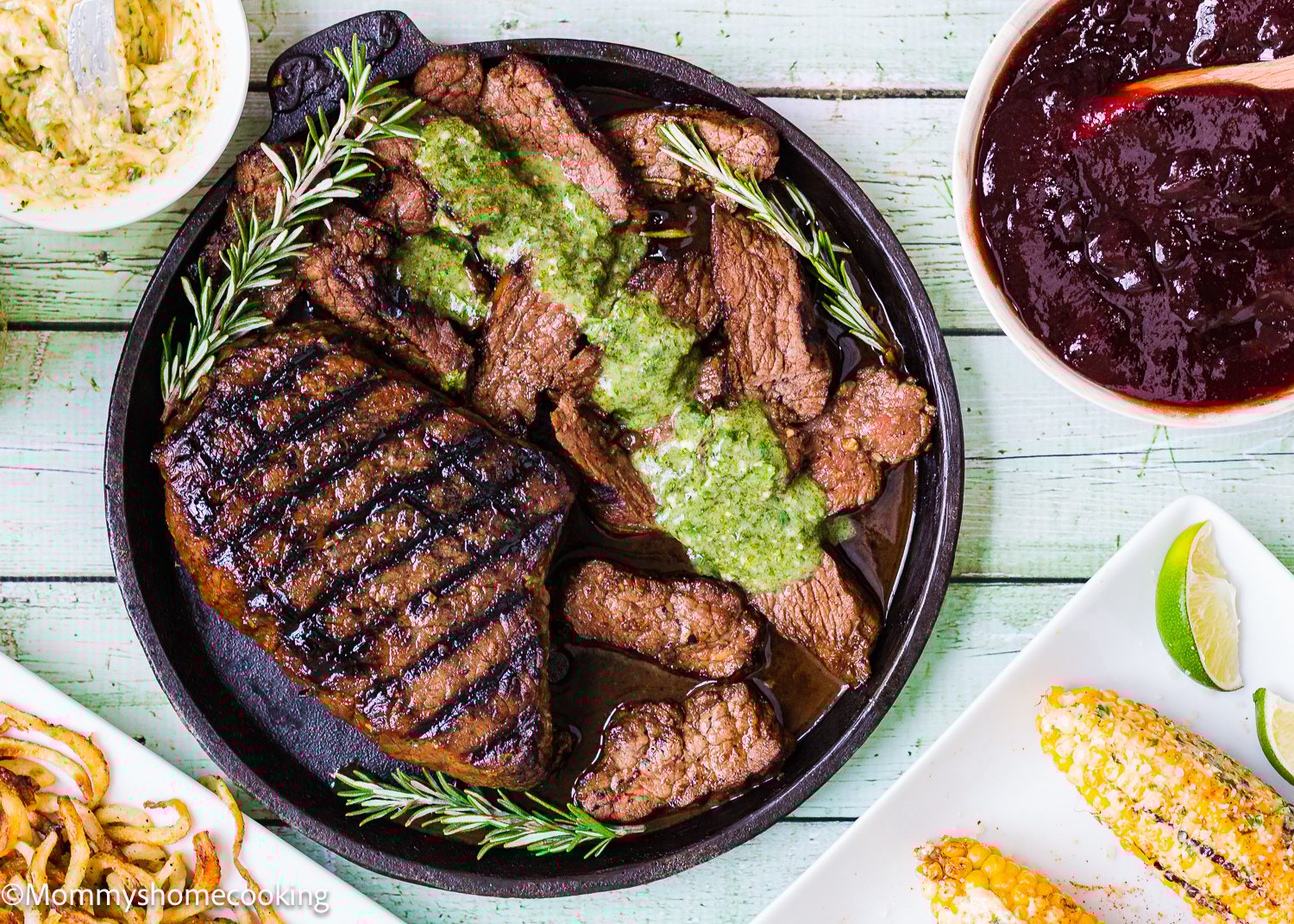 Grilled Balsamic Garlic Steak and beef strips topped with green sauce on a skillet, garnished with rosemary, served with corn, curly fries, and a bowl of reddish sauce.
