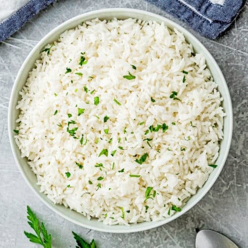 A bowl of cooked white rice garnished with chopped herbs sits on a gray surface beside a sprig of parsley, a spoon, and folded cloth napkins—perfect for showcasing How To Cook Fluffy Rice On Stove (Beginner-Friendly Guide!).