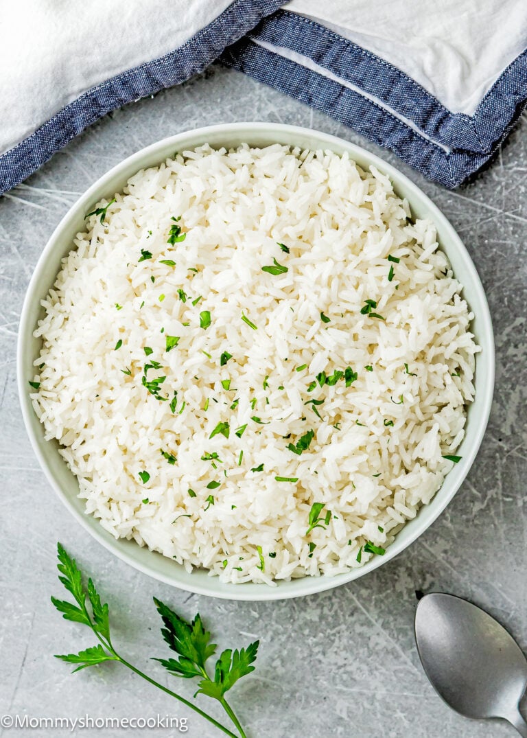 A bowl of cooked white rice garnished with chopped herbs sits on a gray surface beside a sprig of parsley, a spoon, and folded cloth napkins—perfect for showcasing How To Cook Fluffy Rice On Stove (Beginner-Friendly Guide!).