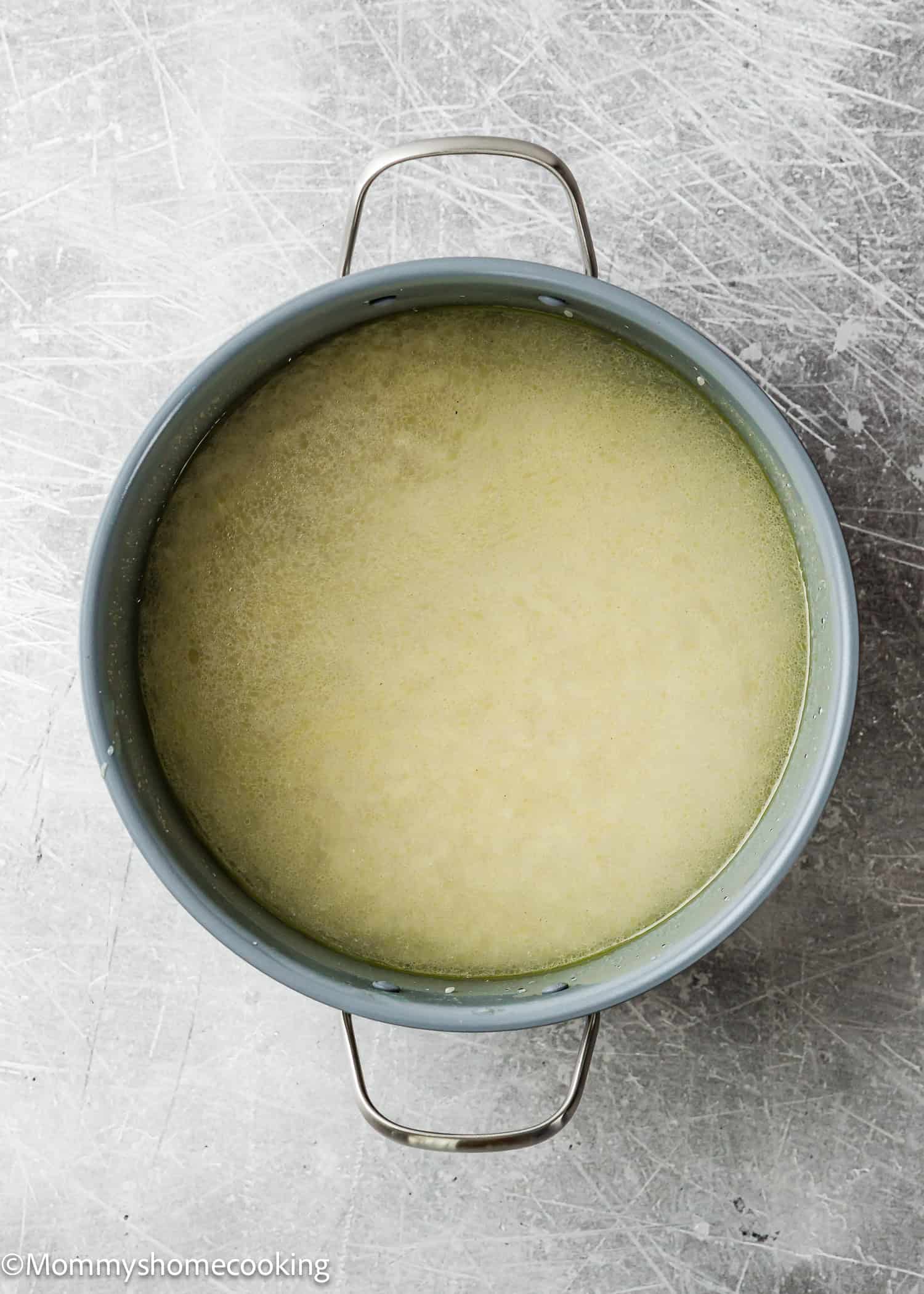A gray pot with a yellowish liquid mixture sits on a scratched metal surface, viewed from above, similar to the first step in How To Cook Fluffy Rice On Stove (Beginner-Friendly Guide!).