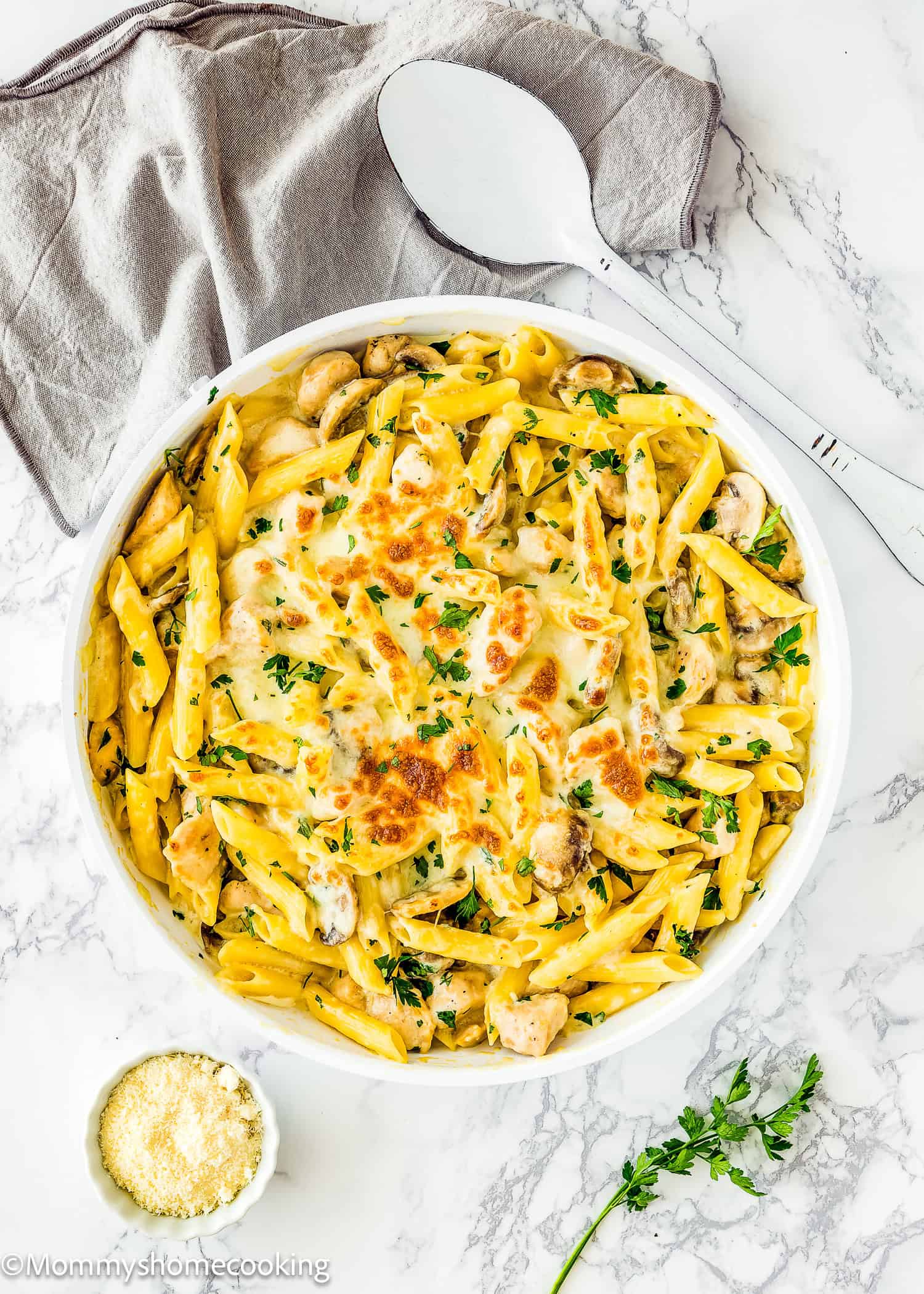 A white dish of One-Pot Easy Chicken Tetrazzini features baked penne pasta, mushrooms, and melted cheese, garnished with parsley. It's accompanied by a gray napkin, a large serving spoon, and a small bowl of grated cheese for added flavor.