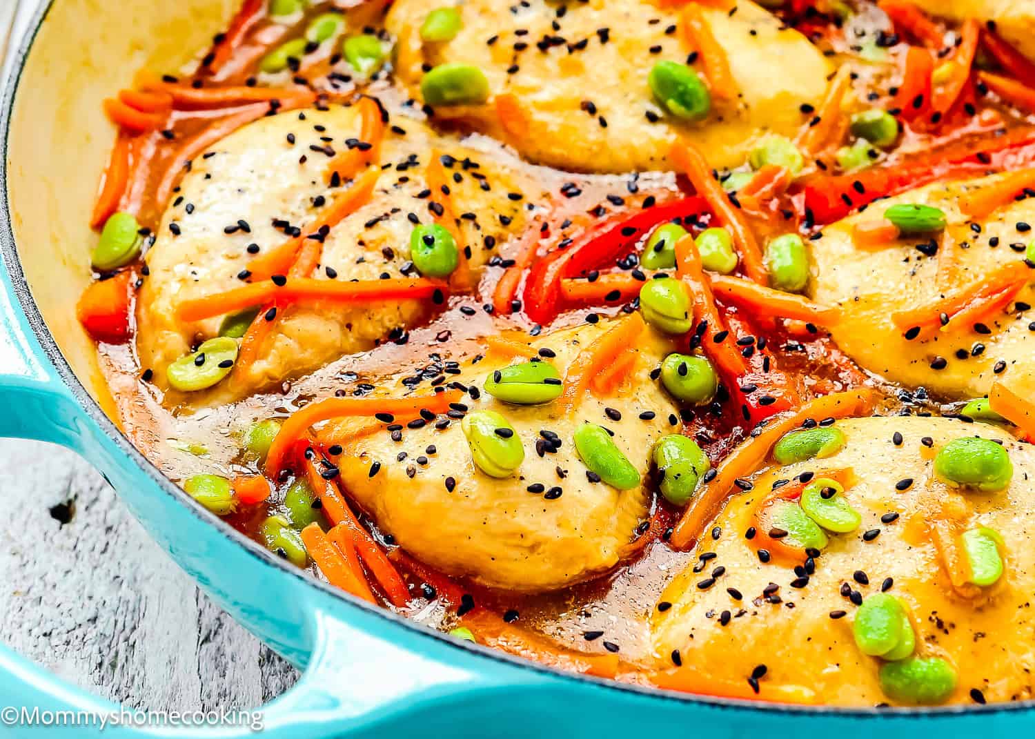 Orange Ginger Chicken breasts in a skillet with a flavorful sauce, topped with black sesame seeds, sliced carrots, red peppers, and edamame.