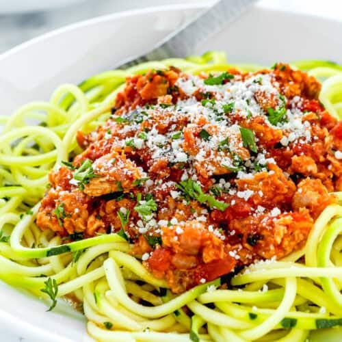 A plate of zucchini noodles topped with a delightful tomato meat sauce and hints of easy tuna ragu, garnished with grated cheese and herbs, with a fork on the side.