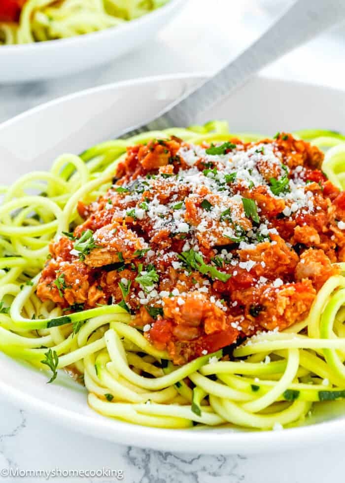 A plate of zucchini noodles topped with a delightful tomato meat sauce and hints of easy tuna ragu, garnished with grated cheese and herbs, with a fork on the side.