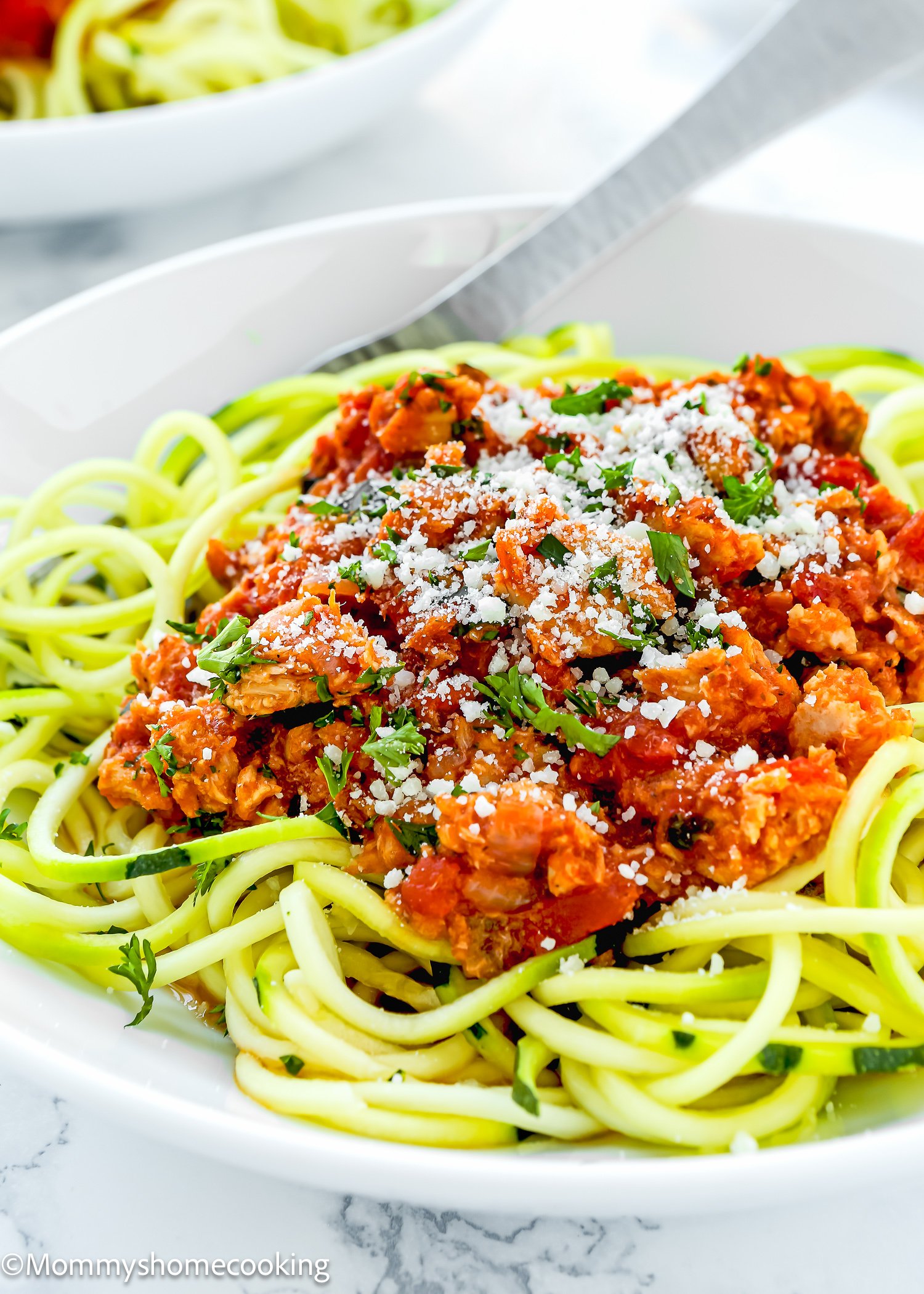 A plate of zucchini noodles topped with a delightful tomato tuna sauce ragu, garnished with grated cheese and herbs, with a fork on the side.