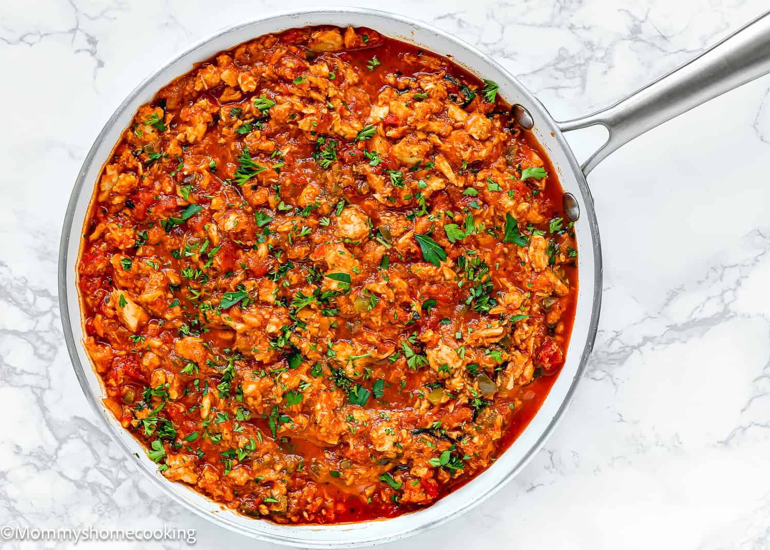 A skillet filled with ground tuna in a red tomato sauce, garnished with chopped herbs on a marble surface.