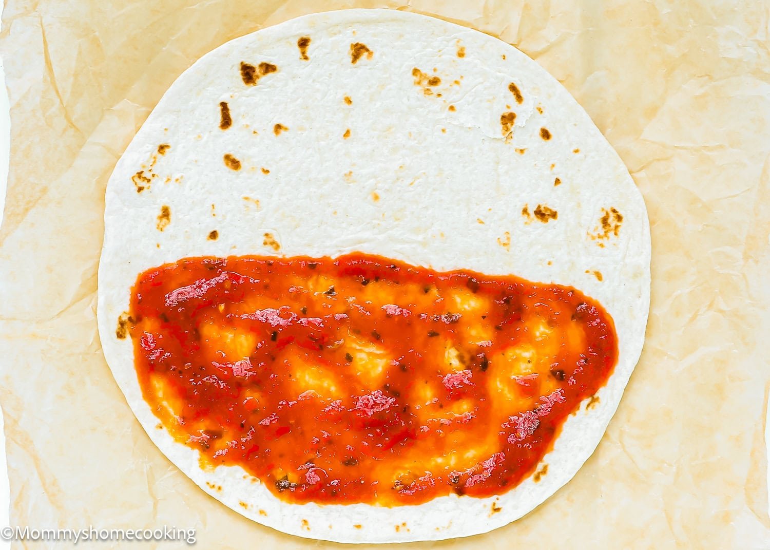A flour tortilla laid flat on parchment paper with pizza sauce spread over the bottom half—Super Easy Pizzadillas ready to be made.