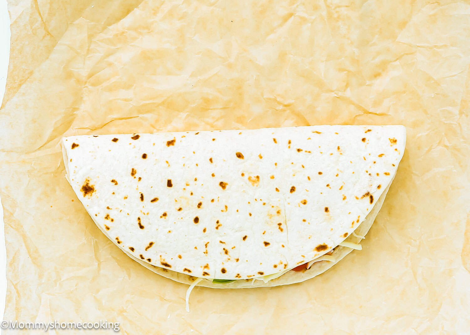 A folded flour tortilla with visible filling underneath, resting on a sheet of brown parchment paper—this is a Super Easy Pizzadillas waiting to be enjoyed.