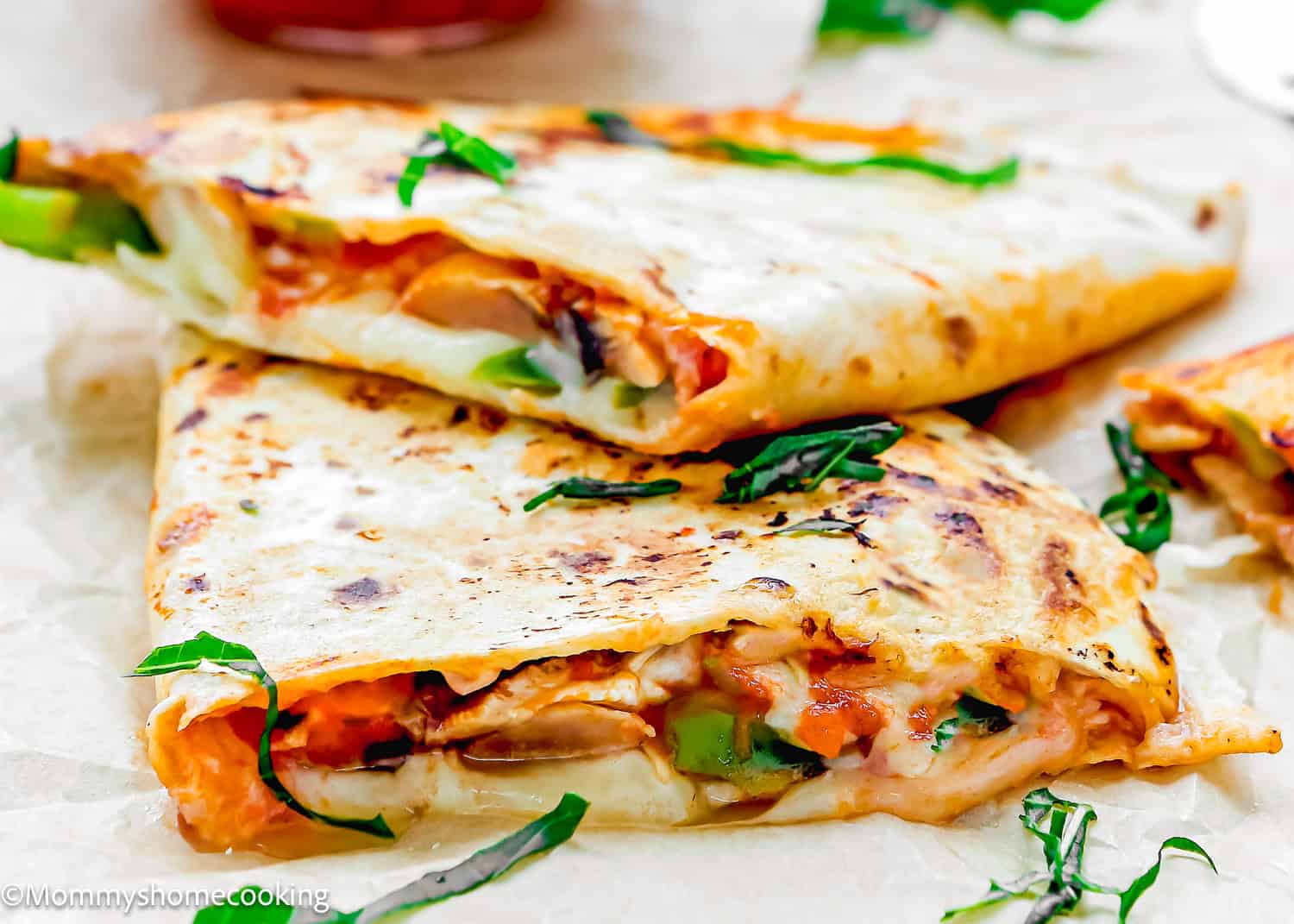 Two folded, golden-brown Super Easy Pizzadillas filled with melted cheese, vegetables, and herbs are stacked on parchment paper.