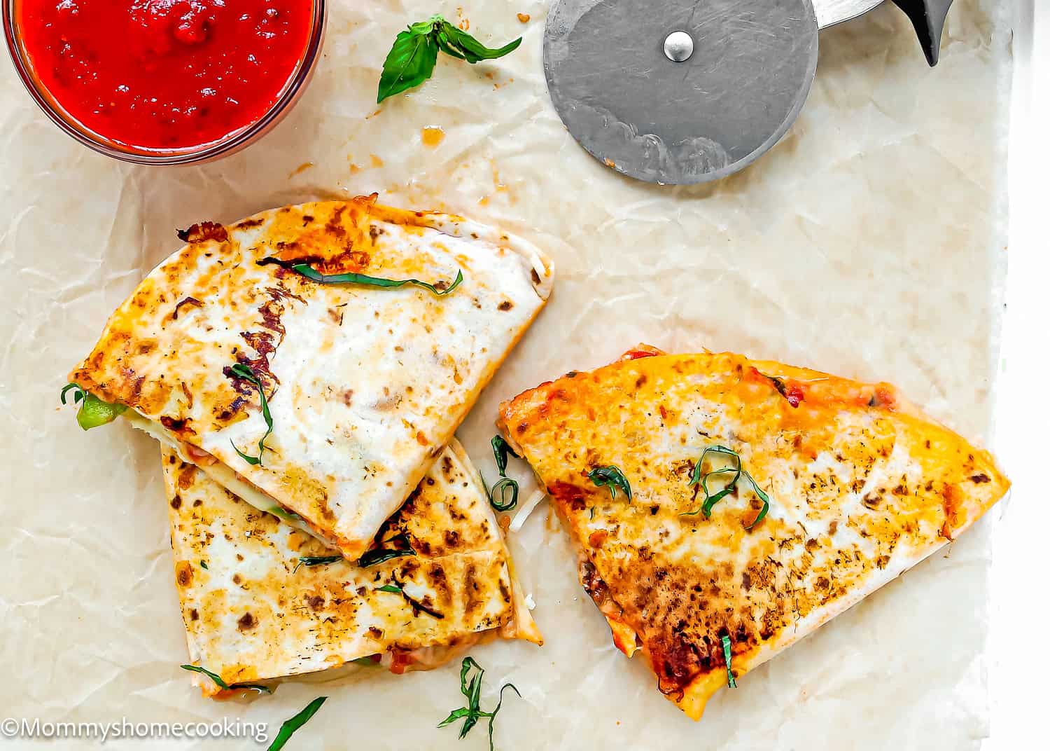 Two folded and grilled quesadillas with melted cheese and herbs, dubbed Super Easy Pizzadillas, served on parchment paper next to a pizza cutter and a bowl of red sauce.