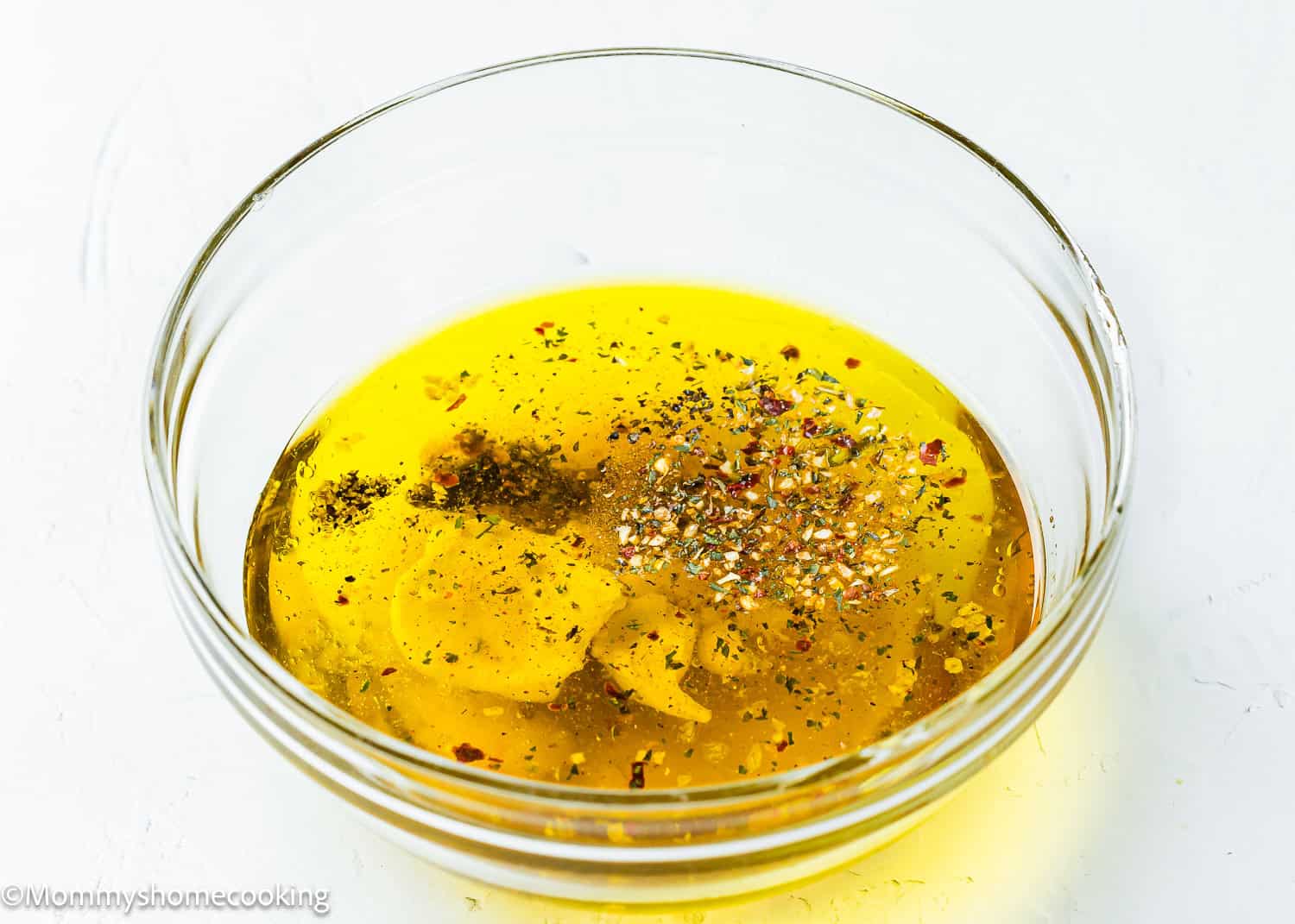 A glass bowl filled with olive oil, garlic slices, black pepper, salt, mixed herbs, and a hint of Apple Cider Honey Dijon Vinaigrette on a white surface.