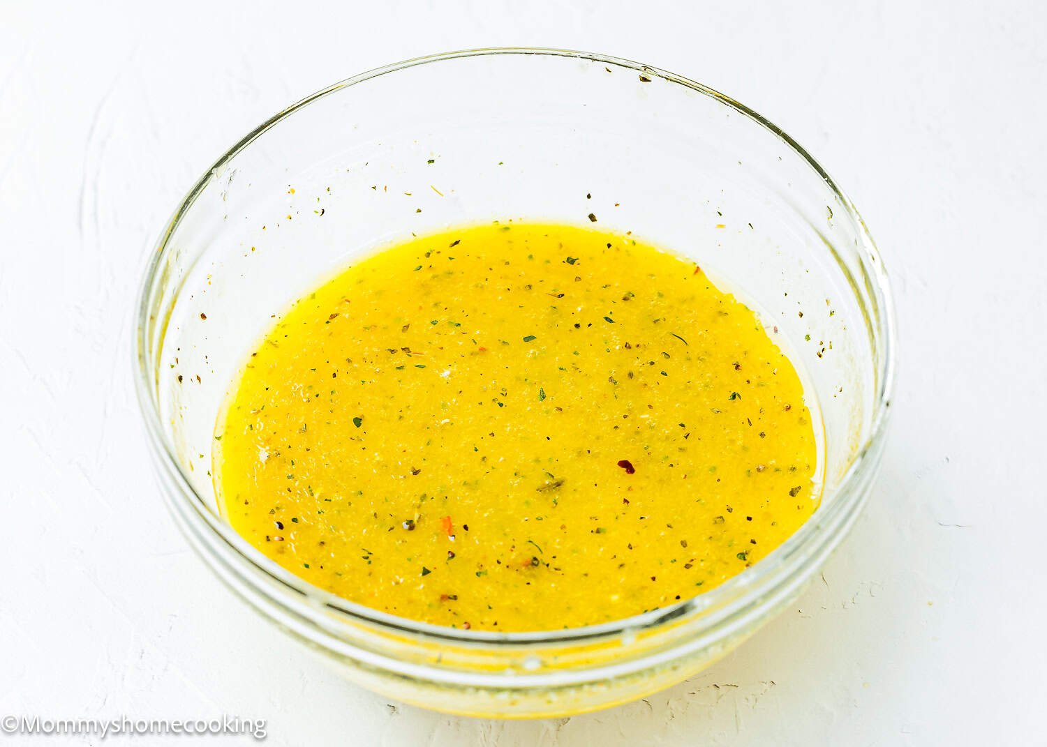 A clear glass bowl filled with Apple Cider Honey Dijon Vinaigrette speckled with herbs, placed on a white surface.
