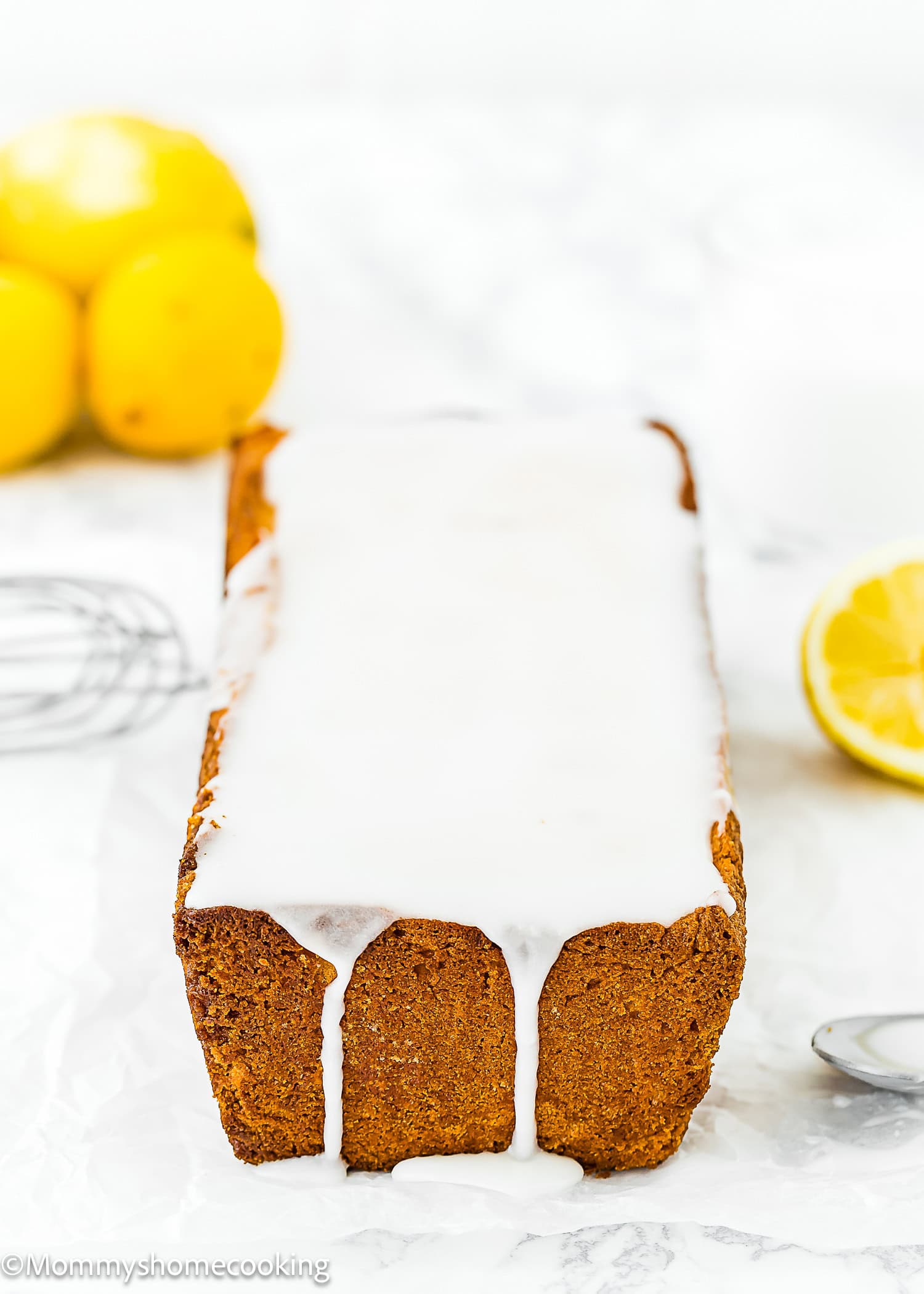 A rectangular loaf of Best Eggless Lemon Pound Cake topped with white icing sits on a white surface, with lemons and a whisk in the background.