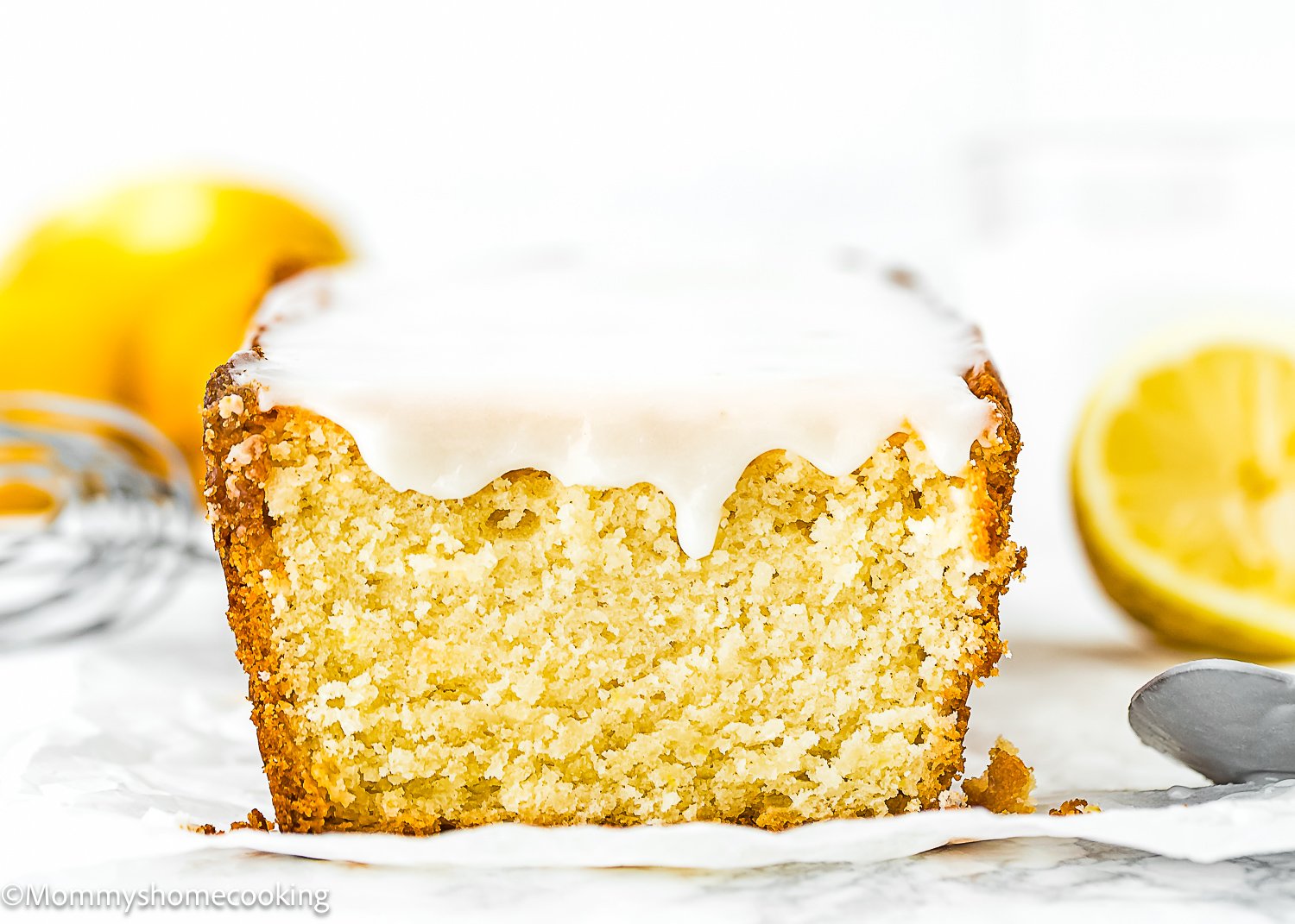 A loaf of Best Eggless Lemon Pound Cake with a thick slice cut off, revealing a moist, dense crumb. A halved lemon and a whisk are visible in the background.