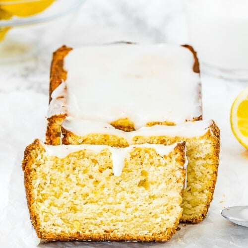 A loaf of Best Eggless Lemon Pound Cake with white glaze, partially sliced, sits on parchment paper. Lemons and a glass of milk are visible in the background.