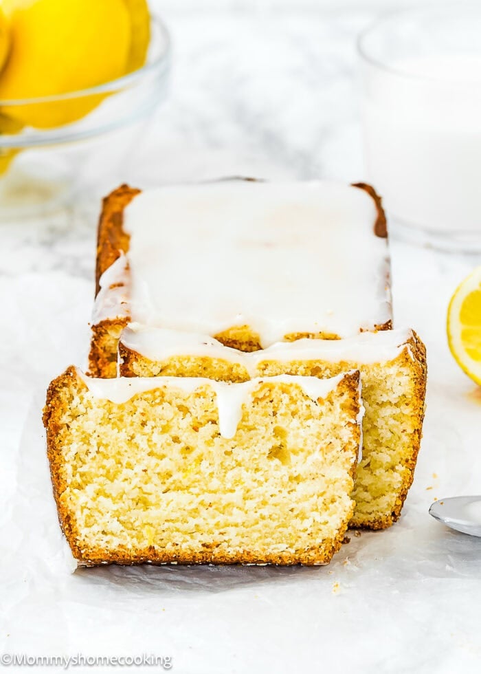 A loaf of Best Eggless Lemon Pound Cake with white glaze, partially sliced, sits on parchment paper. Lemons and a glass of milk are visible in the background.
