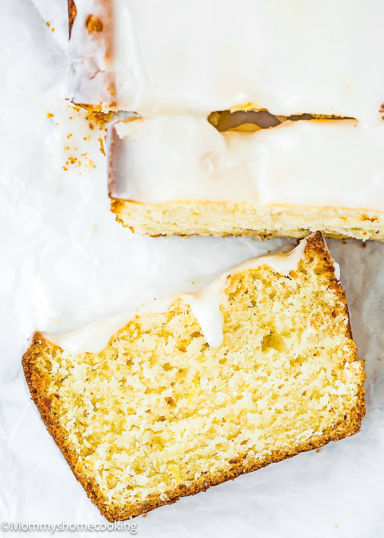 A loaf of the Best Eggless Lemon Pound Cake, partially sliced, with a thick layer of icing on top and crumbs scattered on white parchment paper.