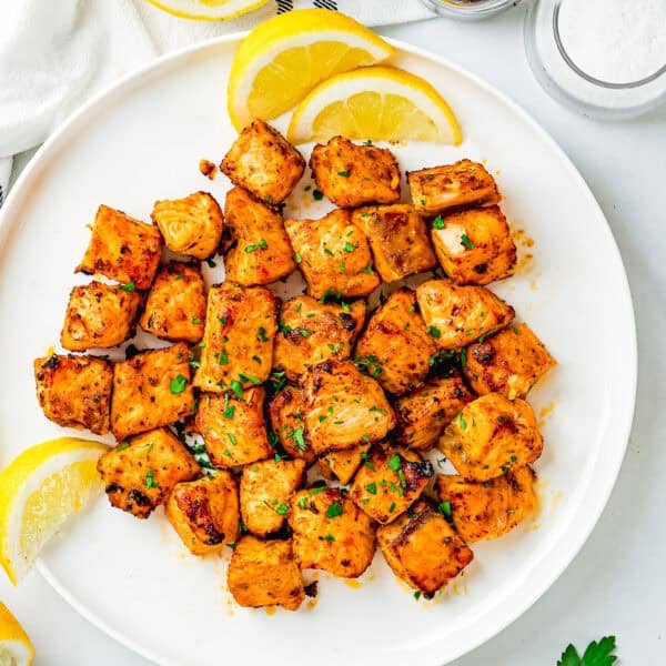 A white plate with Easy Air Fryer Salmon Bites, garnished with chopped herbs and lemon wedges, sits next to jars of salt and pepper.
