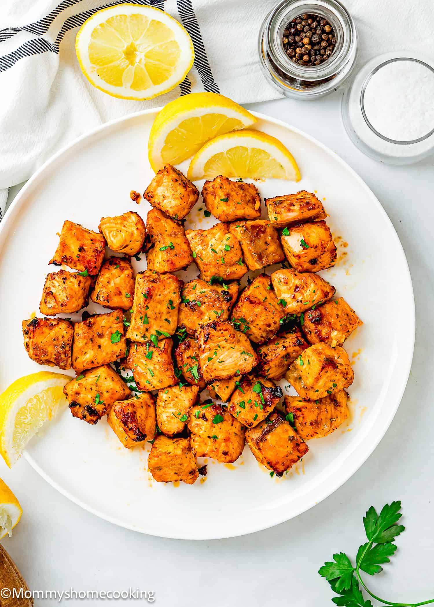 A white plate with Easy Air Fryer Salmon Bites, garnished with chopped herbs and lemon wedges, sits next to jars of salt and pepper.
