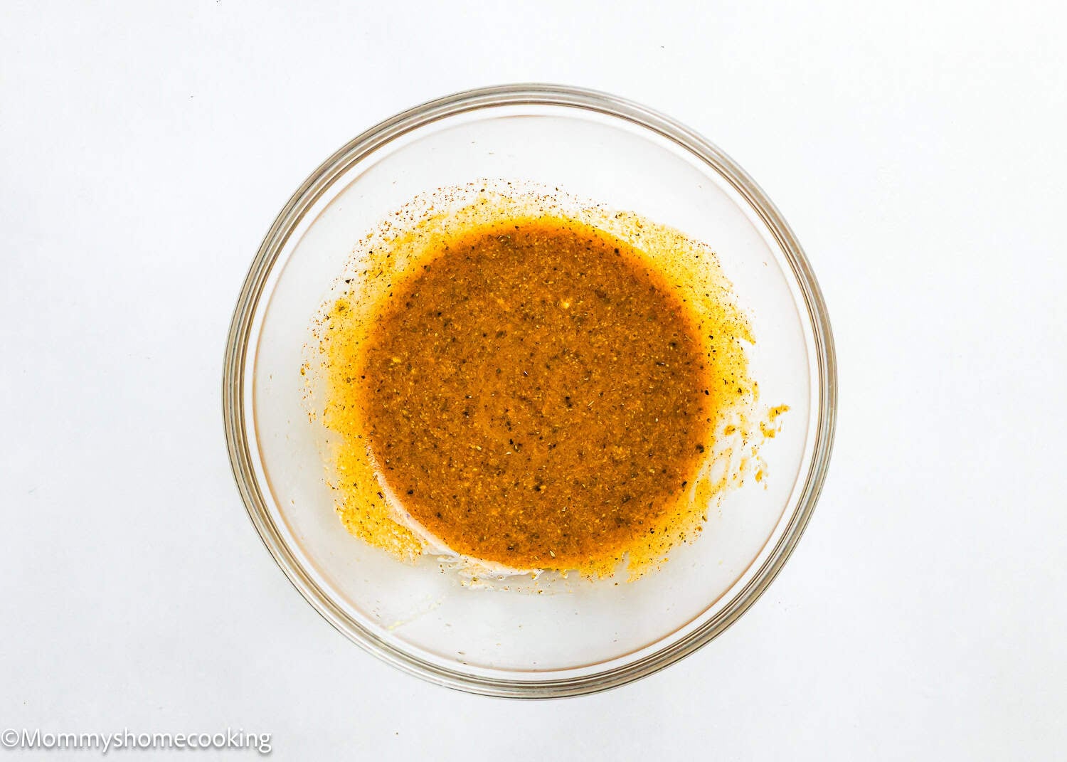 A glass bowl filled with a mixed yellow-orange seasoning or marinade, containing visible specks of herbs and spices for Easy Air Fryer Salmon Bites, on a white background.