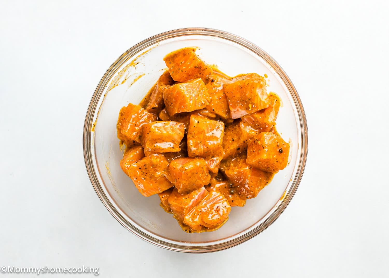 A clear glass bowl contains Easy Air Fryer Salmon Bites—chunks of salmon coated in a seasoned orange marinade, set on a white surface.