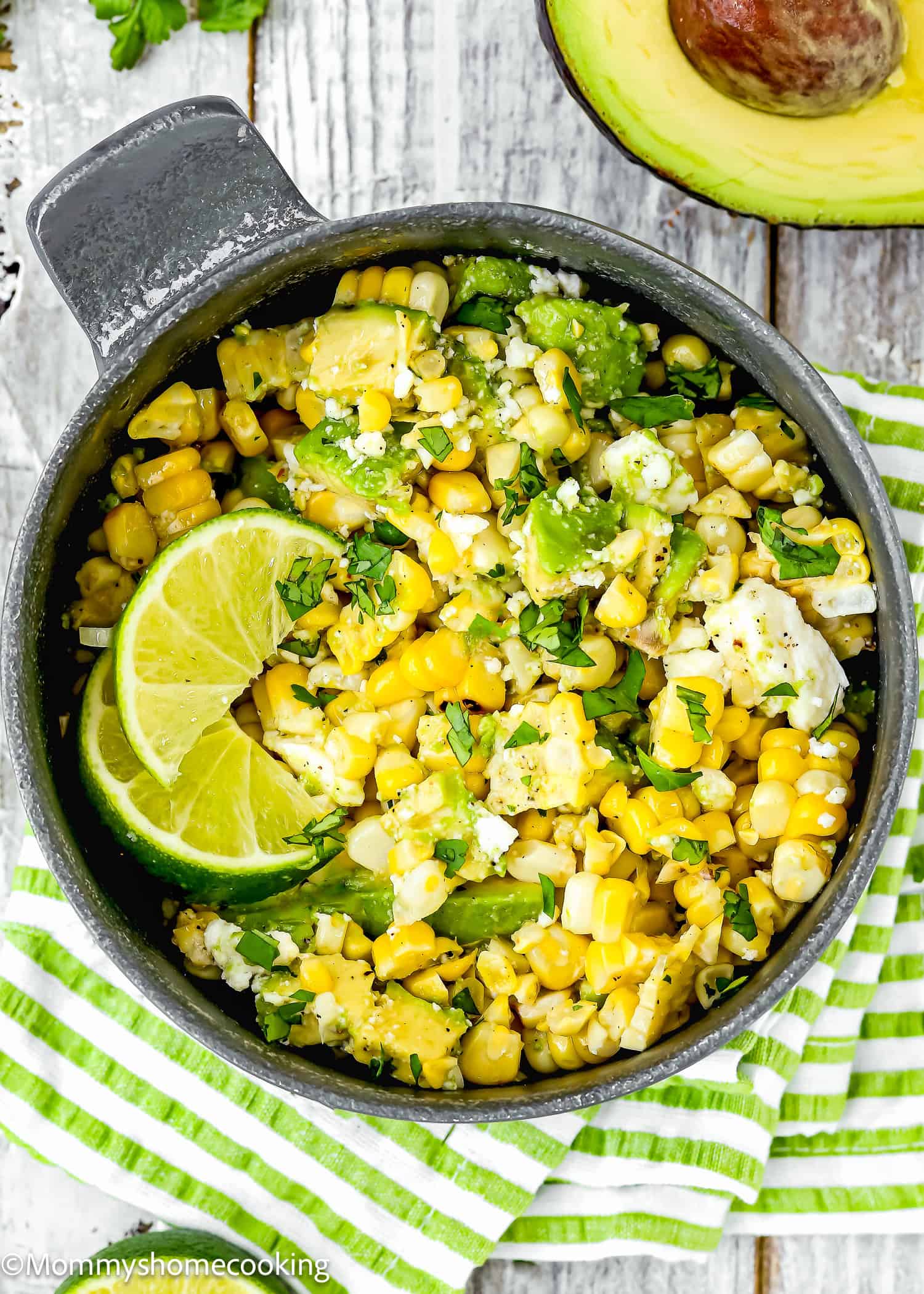 A bowl of Easy Avocado Corn Salad (No Mayo) garnished with lime wedges and chopped herbs sits on a striped kitchen towel next to a halved avocado.