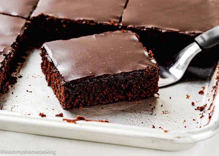 A metal spatula lifts a square piece of Easy Eggless Texas Sheet Cake with glossy frosting from a metal baking pan.