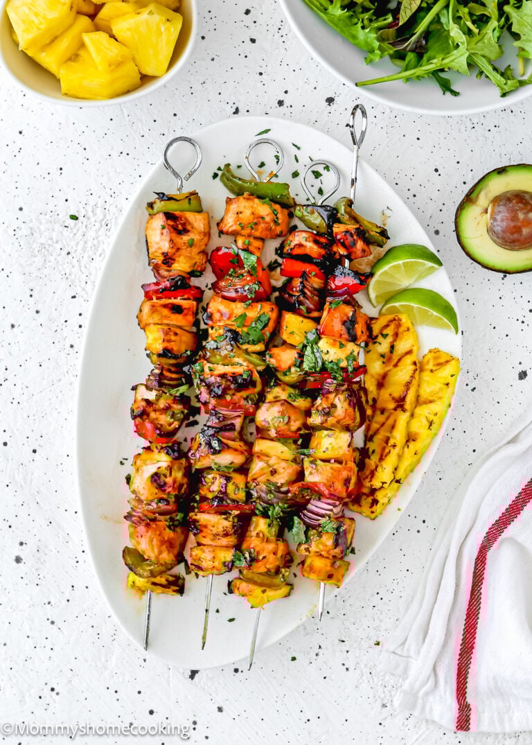 Easy Grilled Hawaiian Chicken Skewers served on a white platter with grilled pineapple slices, lime wedges, and fresh herbs. Bowls of pineapple, arugula, and half an avocado are nearby for a vibrant tropical touch.