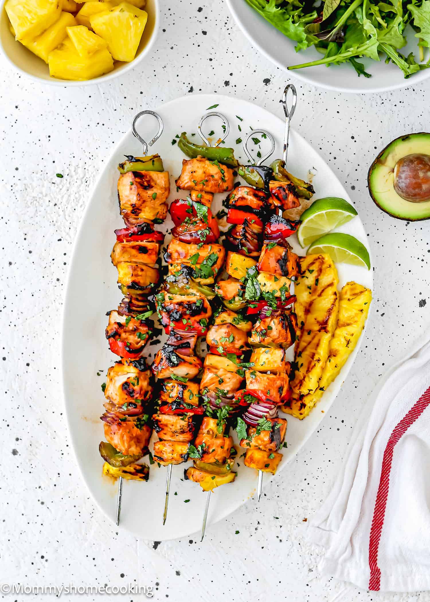 Easy Grilled Hawaiian Chicken Skewers served on a white platter with grilled pineapple slices, lime wedges, and fresh herbs. Bowls of pineapple, arugula, and half an avocado are nearby for a vibrant tropical touch.