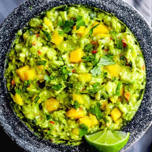 A stone bowl filled with Easy Mango Guacamole, featuring chunks of mango, chopped cilantro, red pepper flakes, and a lime wedge garnish.
