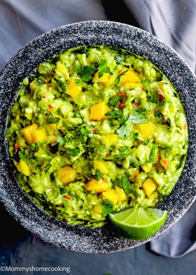 A stone bowl filled with Easy Mango Guacamole, featuring chunks of mango, chopped cilantro, red pepper flakes, and a lime wedge garnish.