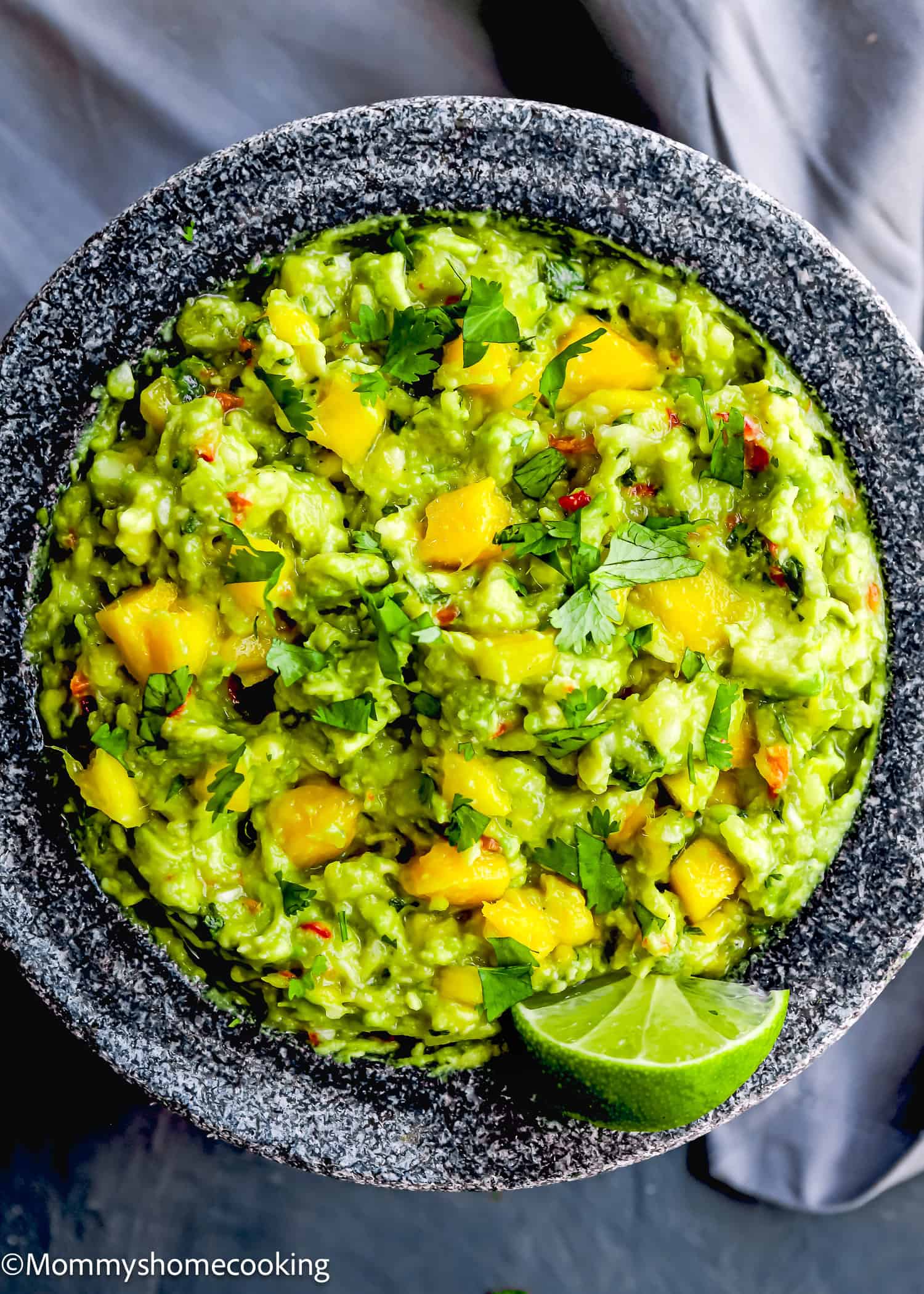 A stone bowl filled with Easy Mango Guacamole, featuring chunks of mango, chopped cilantro, red pepper flakes, and a lime wedge garnish.
