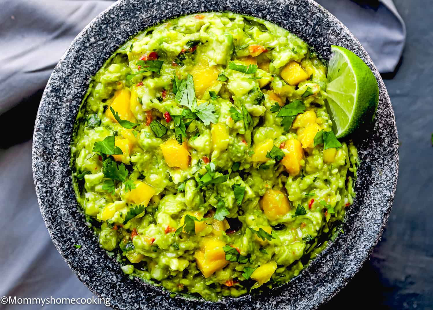 A bowl of Easy Mango Guacamole with chunks of mango, chopped cilantro, and a lime wedge on the side, served in a stone mortar on a gray cloth.