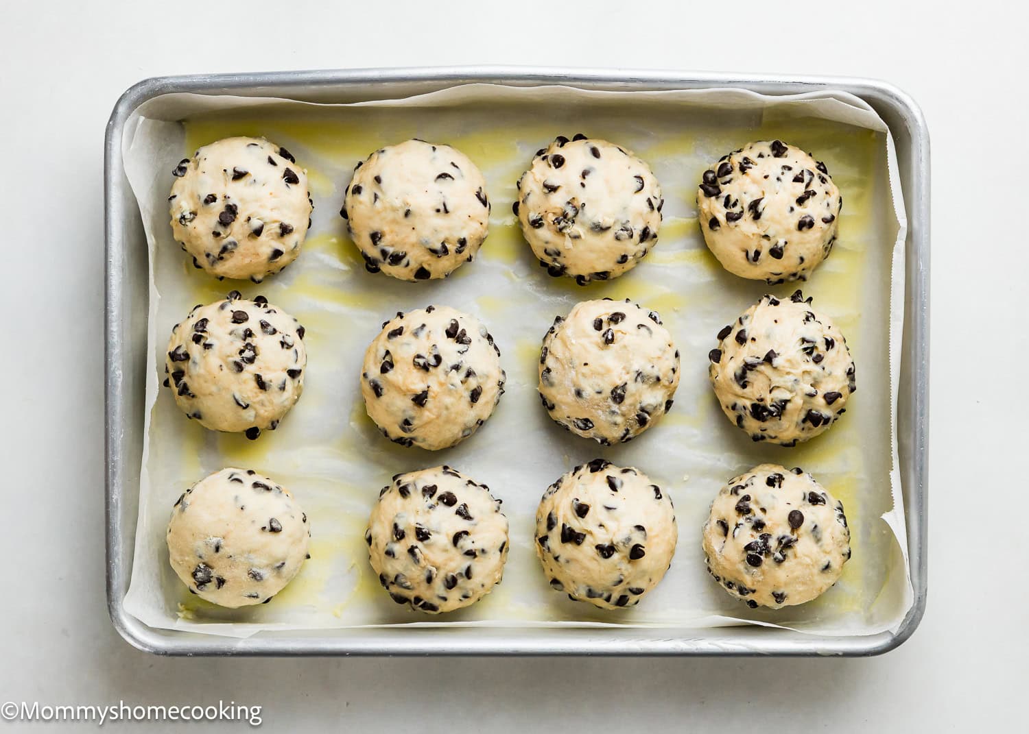 A baking tray lined with parchment paper holds twelve unbaked, round Eggless Chocolate Chip Brioche Rolls mixed with chocolate chips.
