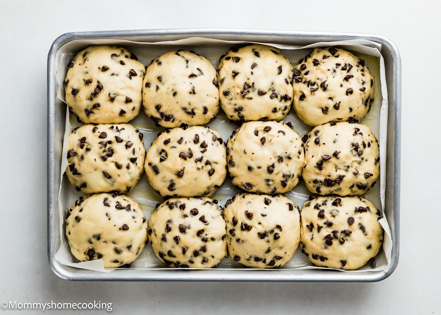 A rectangular baking tray lined with parchment paper holds twelve unbaked Eggless Chocolate Chip Brioche Rolls, each dough ball studded with chocolate chips and arranged in three neat rows.