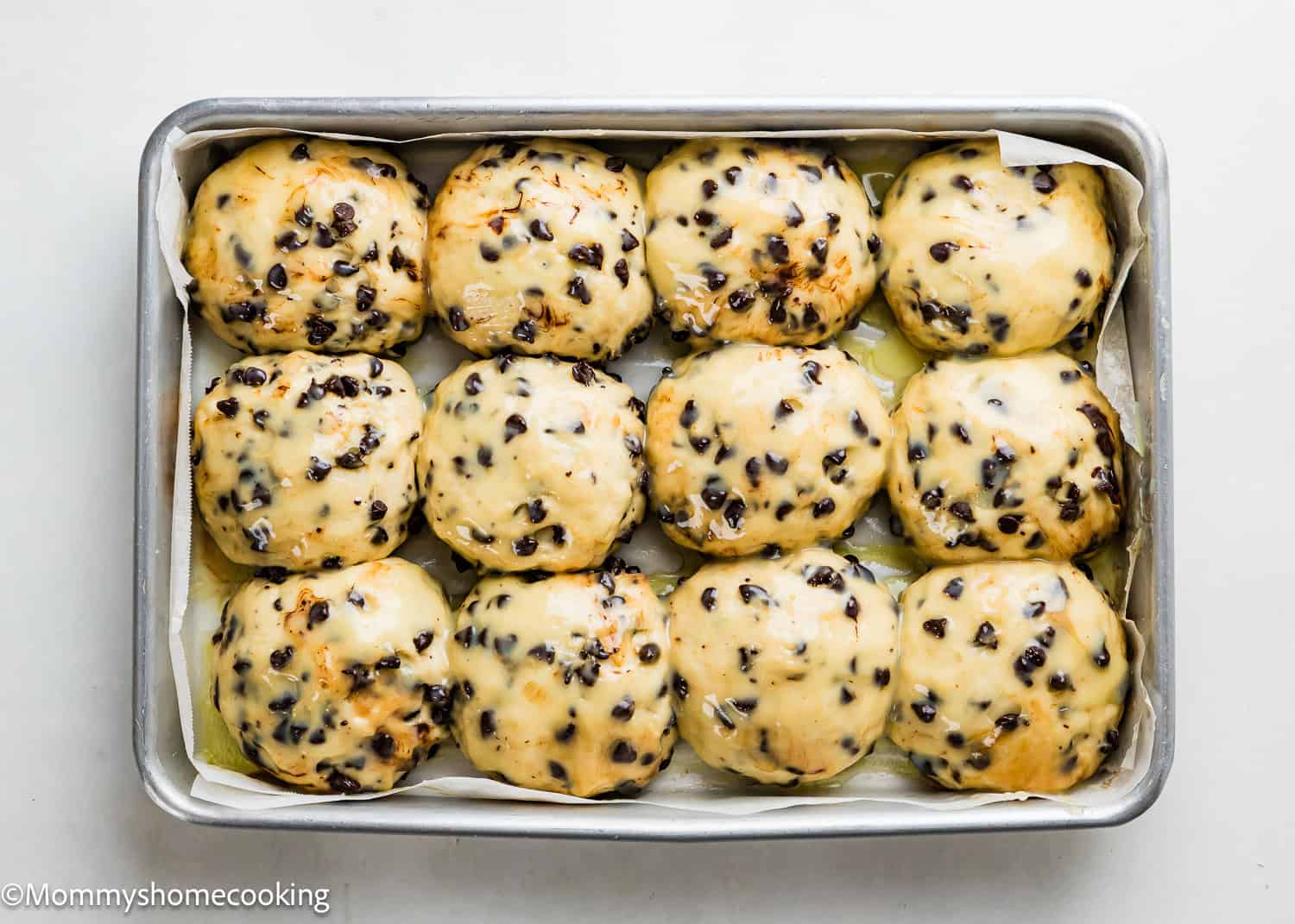 A baking tray filled with twelve Eggless Chocolate Chip Brioche Rolls, their glazed tops studded with chocolate chips, arranged in three rows on parchment paper.