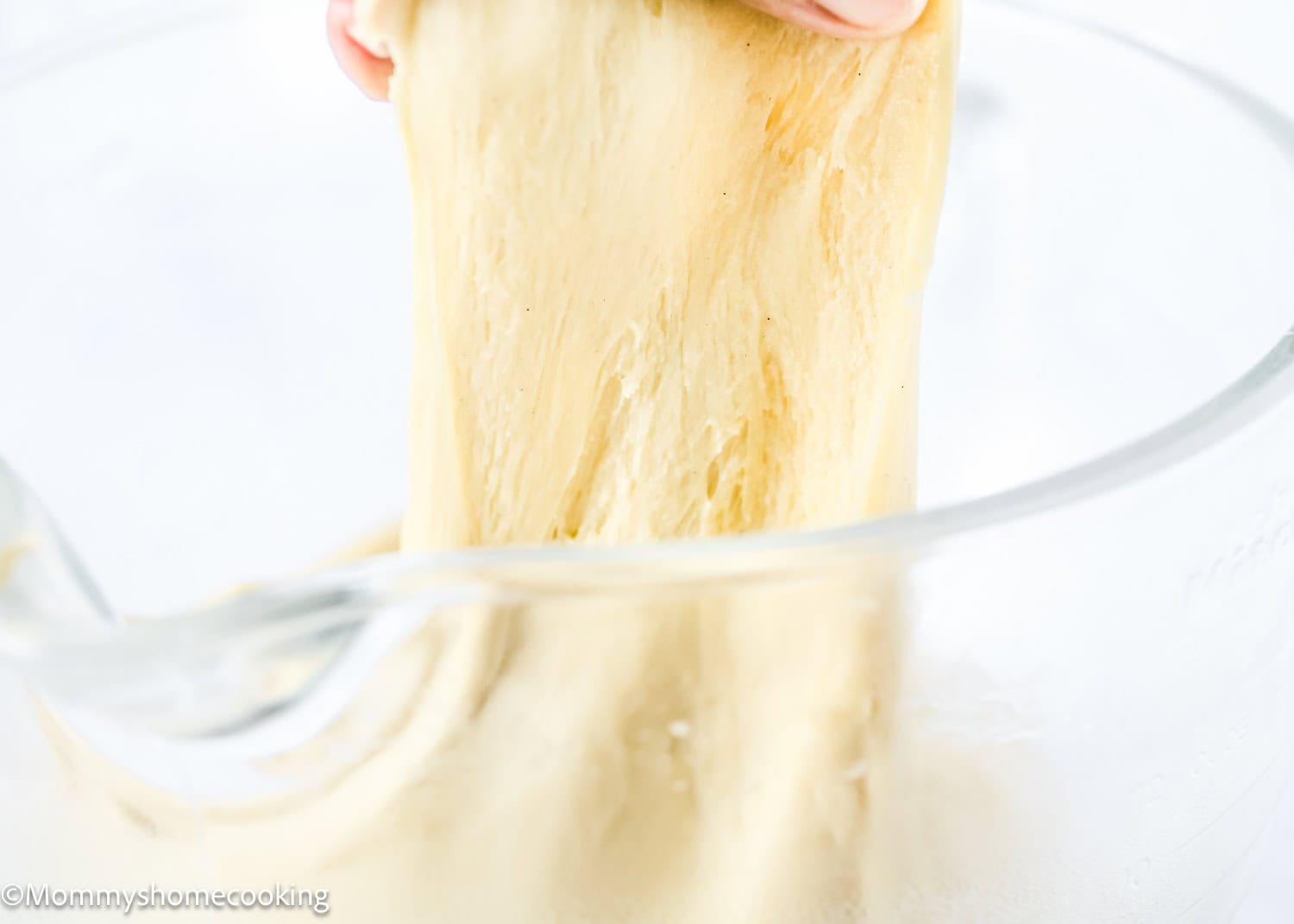 A hand stretches a soft, elastic dough for Eggless Chocolate Chip Brioche Rolls inside a clear glass mixing bowl.