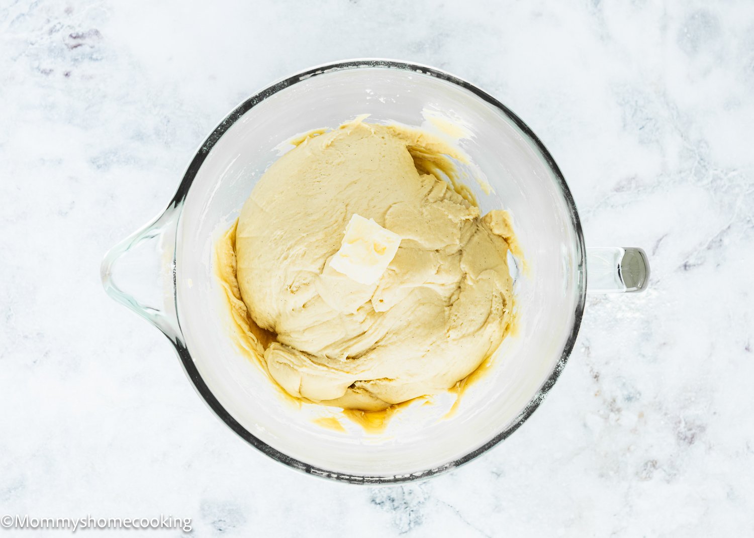 Glass bowl containing dough for Eggless Chocolate Chip Brioche Rolls with a pat of butter on top, placed on a white marble surface.