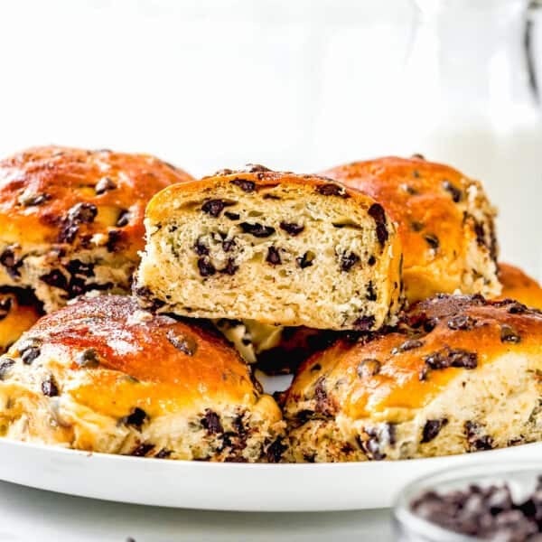 A plate of golden brown Eggless Chocolate Chip Brioche Rolls, with one bun cut in half to reveal the chocolate chip filling, sits beside a bowl of chocolate chips and a glass of milk in the background.
