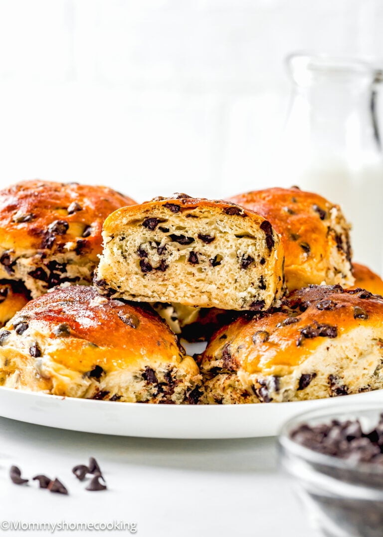 A plate of golden brown Eggless Chocolate Chip Brioche Rolls, with one bun cut in half to reveal the chocolate chip filling, sits beside a bowl of chocolate chips and a glass of milk in the background.