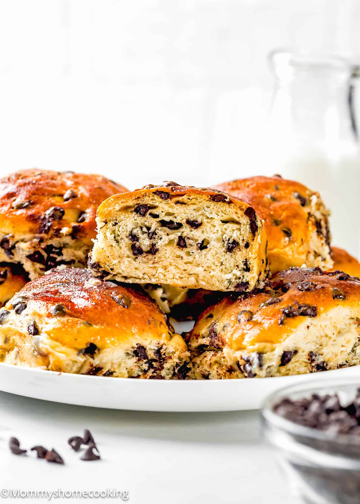 A plate of golden brown Eggless Chocolate Chip Brioche Rolls, with one bun cut in half to reveal the chocolate chip filling, sits beside a bowl of chocolate chips and a glass of milk in the background.