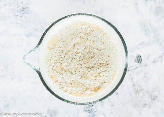 A glass mixing bowl filled with flour sits on a white marble surface, ready to make Eggless Chocolate Chip Brioche Rolls.