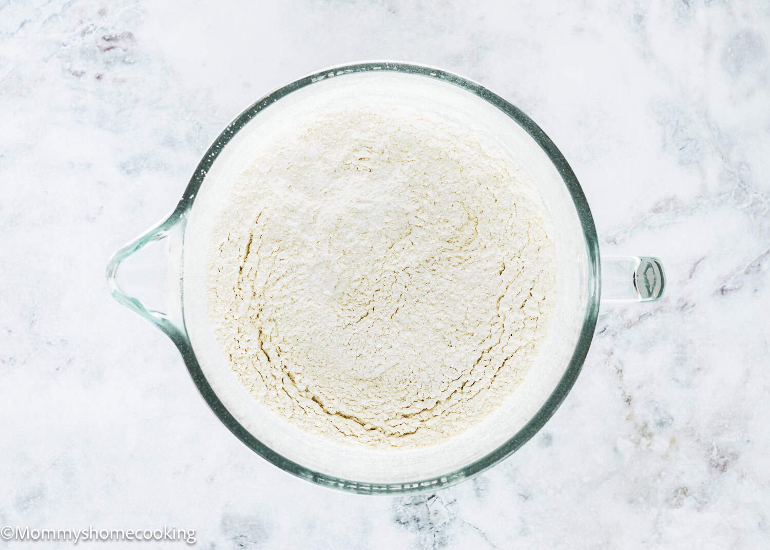 A glass mixing bowl filled with flour sits on a white marble surface, ready to make Eggless Chocolate Chip Brioche Rolls.