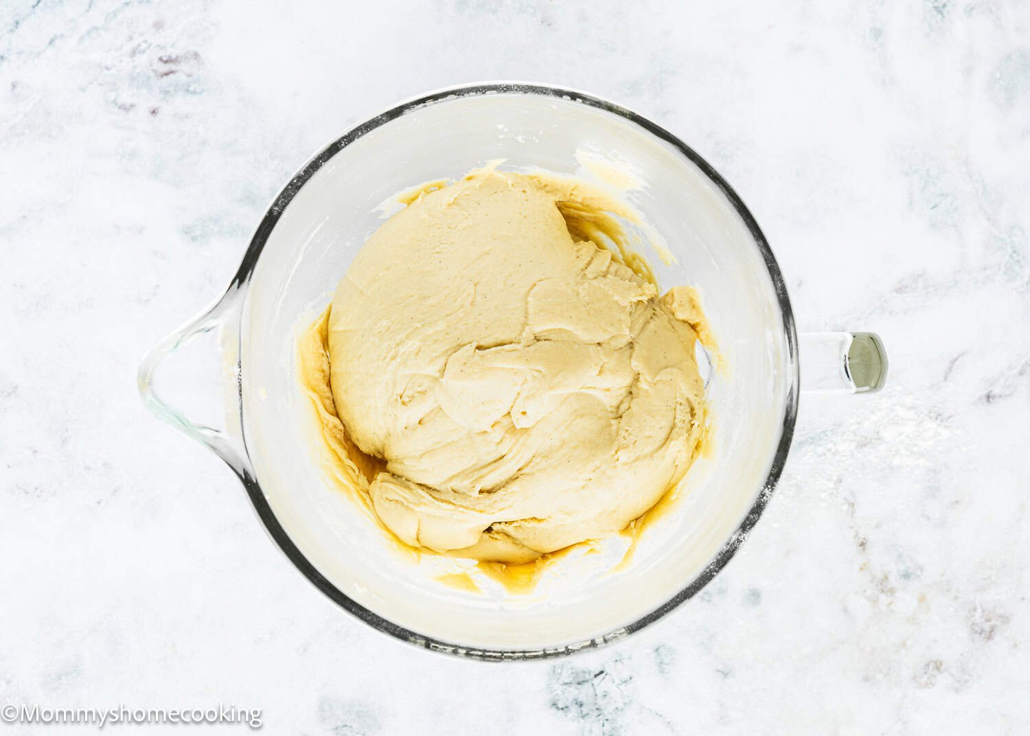 A glass mixing bowl containing a ball of Eggless Chocolate Chip Brioche Rolls dough sits on a marble countertop.